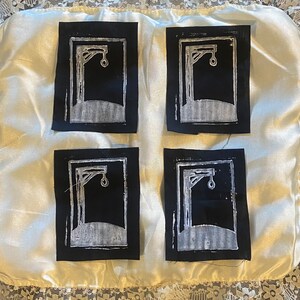 May include: Four black fabric squares with white embroidered images of a gallows with a noose. The squares are arranged in a 2x2 grid on a white satin background.