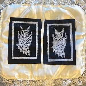 May include: Two black and white pillowcases with a white bat design on each. The pillowcases are on a white satin pillow with a white lace trim.