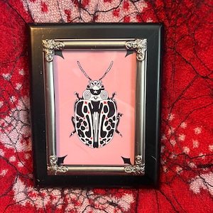 Puede incluir: Una impresión artística enmarcada con un diseño estilizado de escarabajo en blanco y negro con detalles rojos sobre un fondo rosa. El marco negro tiene esquinas decorativas plateadas. La obra se exhibe sobre un fondo rojo con una superposición de encaje negro.