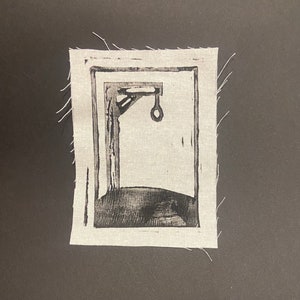 May include: A black and white print of a gallows with a noose hanging from it. The print is framed in a white fabric with a frayed edge.