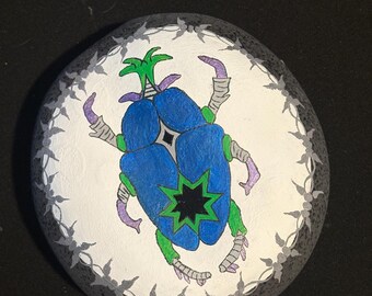 May include: Hand-painted rock with a blue and green beetle design. The beetle has a star-shaped body and is surrounded by a gray border.