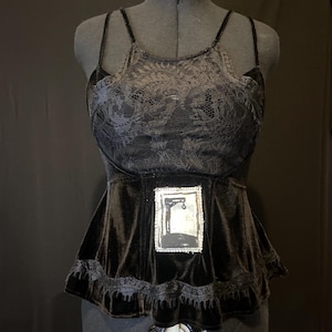 May include: A black velvet top with a lace overlay and a small framed image of a hanging figure. The top has a ruffled hem.