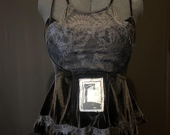 Pode incluir: Um top de veludo preto com uma sobreposição de renda e uma pequena imagem emoldurada de uma figura pendurada. O top tem uma bainha com babados.