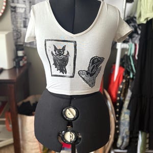 May include: White V-neck crop top with two black graphic prints. One print is a bat in a frame, the other is a hand with a design. The top is displayed on a black dress form.