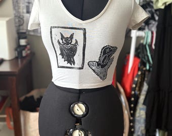 Pode incluir: Top curto branco com decote em V com duas estampas gráficas pretas. Uma estampa é um morcego em uma moldura, a outra é uma mão com um desenho. O top é exibido em um manequim preto.