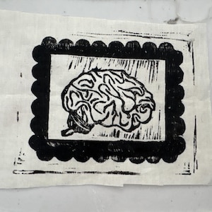 May include: A black and white linocut print of a brain on a square of fabric. The brain image is framed by a black scalloped border. The fabric is a light beige color.