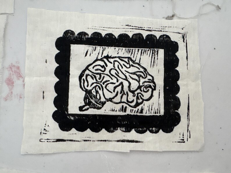 May include: A black and white linocut print of a brain on a square of fabric. The brain image is framed by a black scalloped border. The fabric is a light beige color.