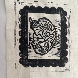 May include: A black and white linocut print on a square of off-white fabric. The print features a stylized, organic shape within a scalloped rectangular border. The fabric has a slightly textured appearance.