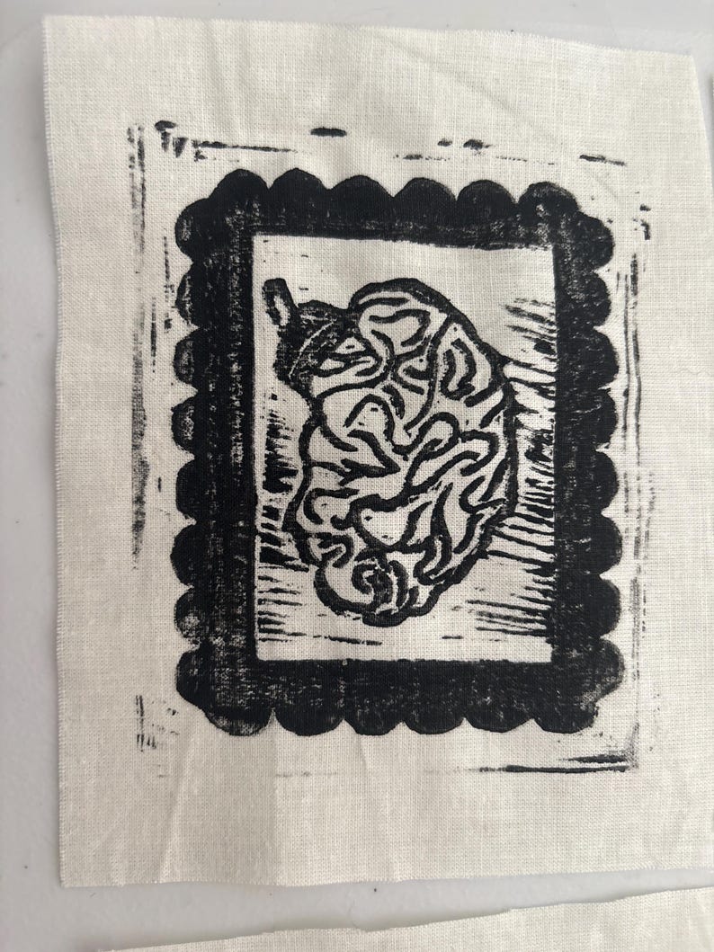 May include: A black and white linocut print on a square of off-white fabric. The print features a stylized, organic shape within a scalloped rectangular border. The fabric has a slightly textured appearance.