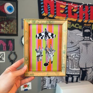 May include: A framed art print featuring a whimsical carousel scene with two clown figures in black and white. The background has vertical stripes in pink, green, and orange. The frame is gold-colored.