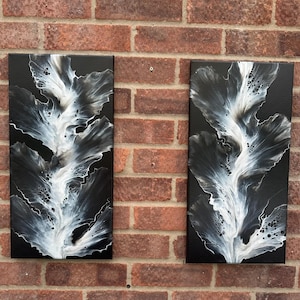 May include: Two abstract paintings with a black background and white and grey floral designs. The artwork is displayed on a brick wall. The paintings are rectangular and vertically oriented.