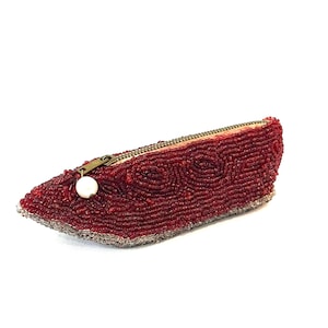 May include: A beaded red clutch with a zipper closure and a pearl-like charm. The clutch is covered in small, dark red beads, with a decorative pattern on the front. The zipper is a brass color, and the edges are lined with silver beads.