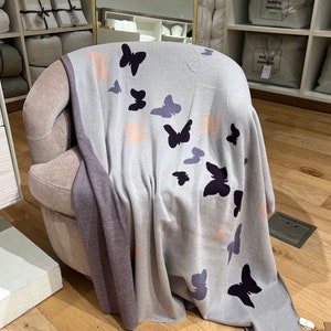 May include: A soft, gray throw blanket with a subtle ombre effect and a pattern of dark purple butterflies. The blanket is draped over a plush, light pink armchair.