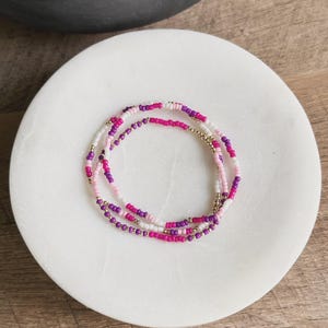 May include: A multi-strand beaded necklace with white, pink, and purple beads. The necklace is made of small, round beads and has a gold clasp.