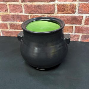 May include: A round ceramic pot with a matte black exterior and a vibrant lime green interior. The vessel features two small, protruding side handles and a wide, open top, showcasing a smooth, rounded silhouette and a sturdy base.