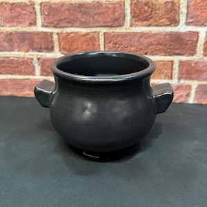 May include: A matte black ceramic pot with a rounded body, a wide circular opening, and two small side handles. The vessel features a smooth, dark finish and a sturdy base, presenting a simple, functional design for various household uses.