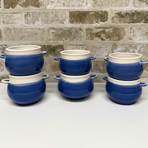 Puede incluir: Seis cuencos de sopa de cerámica con un diseño de dos tonos. Los cuencos son azules con un borde y asas blancos. Los cuencos están apilados en pares sobre una superficie blanca, con una pared de ladrillos al fondo.