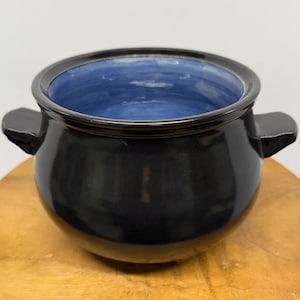 May include: A black ceramic bowl with a glossy finish and two side handles. The interior is a vibrant blue, contrasting with the dark exterior. The bowl sits on a wooden surface, showcasing its shape and design.