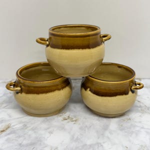 Puede incluir: Tres cuencos de cerámica para sopa con asas. Los cuencos son de color beige claro con una banda marrón oscuro alrededor de la parte superior. Los cuencos están apilados uno encima del otro. Los cuencos tienen un aspecto rústico y hecho a mano.