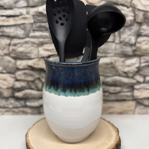 Op de afbeelding: Een keramische keukengerei houder met een witte basis, een turquoise middengedeelte en een donkerblauwe bovenkant. Zwarte keukengerei, waaronder een sleuflepel en een pollepel, zitten erin. De houder staat op een houten schijf.