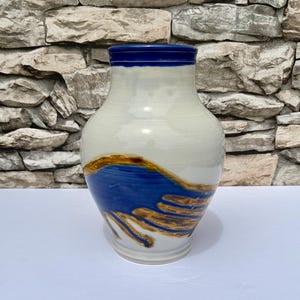 Könnte beinhalten: Eine Keramikvase mit cremefarbenem Körper und blauem Rand. Die Vase hat ein blaues und braunes abstraktes Design auf der unteren Hälfte. Die Vase ist etwa 25 cm hoch.