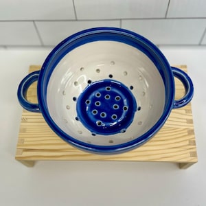 Könnte beinhalten: Ein Keramik-Gemüsesieb mit weißem Innenraum und einem leuchtend blauen Rand und Boden. Das Sieb hat zwei Schlaufengriffe und zahlreiche Abflusslöcher. Es steht auf einem kleinen Holzständer und zeigt sein handwerkliches Design.