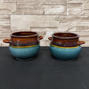 Pode incluir: Duas tigelas de sopa de cerâmica com alças. As tigelas têm um esmalte de duas cores, com a parte superior marrom e a base azul turquesa. As tigelas são redondas e têm pequenas alças em ambos os lados. As tigelas estão sobre uma superfície preta.