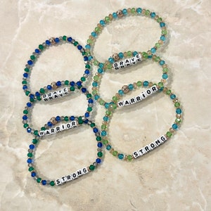 May include: Four beaded bracelets with the words "Brave", "Warrior", and "Strong" spelled out in white letters on black tiles. The bracelets are made with blue, green, and gold beads.