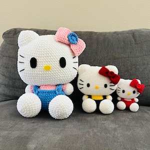 Puede incluir: Tres peluches de Hello Kitty de crochet. La más grande es blanca con un lazo rosa y ojos azules. La del medio es blanca con un lazo rojo y un mono amarillo. La más pequeña es blanca con un lazo rojo y un mono rojo.
