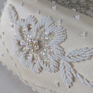 Könnte beinhalten: Eine weiße Satin-Clutch mit einem perlenbesetzten Blumenmuster. Die Blume besteht aus weißen Perlen und schimmernden Pailletten.