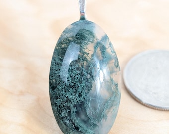 May include: Oval-shaped pendant with a silver bail. The pendant features a translucent stone with green, moss-like inclusions. The stone is set against a light-colored wooden surface. A silver coin is visible in the background.