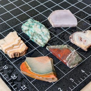 May include: Assortment of polished, raw gemstones in various colors, including green, red, orange, and beige. The stones are displayed on a black grid surface with measurement markings.