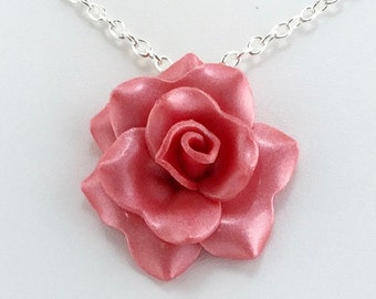 May include: A rose pendant necklace. The pendant is a three-dimensional rose in a metallic pink color. The necklace chain is silver-colored and connects to the rose pendant.