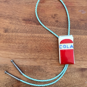 May include: A bolo tie featuring a rectangular pendant with a white background, a red top arch, a red bottom rectangle, and the word COLA in blue letters. The pendant is attached to a braided light blue cord with silver-toned metal tips at the ends.