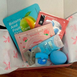 Könnte beinhalten: Geschenkbox mit Hautpflegeprodukten. Enthält eine blaue Gesichtsmaske mit dem Text "Botany And Fruits Skin Care", eine Lippenmaske mit Pfirsich, ein blaues Peeling, ein kleines Glas Vaseline und eine blaue Badekugel. Die Box ist mit rosa und weißem Papier ausgekleidet.