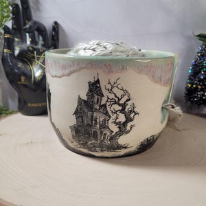 May include: A ceramic yarn bowl with a cream-colored base and a light pinkish-grey glazed rim. The exterior features a black ink illustration of a Victorian-style haunted house and a gnarled, leafless tree. A yarn guide notch is visible on the side.