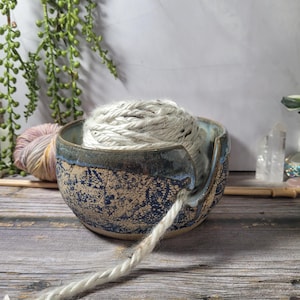 May include: A round ceramic yarn bowl featuring a dark blue and tan lace-like pattern on the exterior with a smooth blue glazed rim. A deep notch is cut into the side for yarn to pass through, and a ball of thick, cream-colored yarn sits inside.