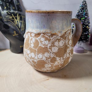May include: A ceramic mug with a speckled tan base featuring a white skull pattern. The upper portion of the mug has a smooth, light blue and lavender glaze that drips down over the textured, patterned lower section. It includes a sturdy handle.