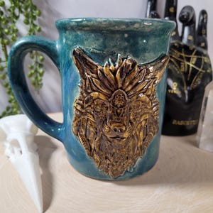 May include: A teal ceramic mug with a detailed, raised relief of a wolf's head in shades of brown and gold. The mug has a rounded shape with a sturdy handle. The background is blurred, suggesting a natural or mystical theme.