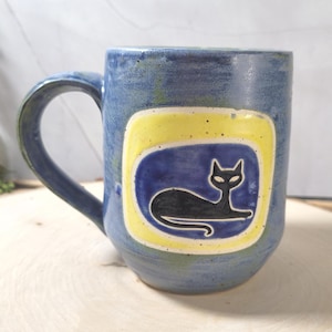 May include: Blue ceramic mug with a black cat design. The cat is inside a blue rectangle with a yellow border. The mug has a curved handle and is on a wooden surface. The mug is a unique piece of art.