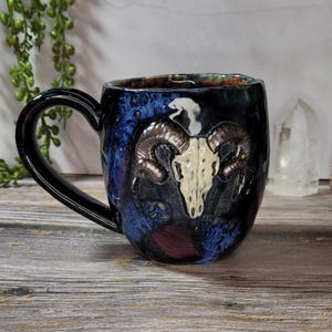 May include: A ceramic mug with a black handle and a dark blue, black, and maroon exterior. The mug features a white ram skull design with brown horns. The mug is on a wooden surface.