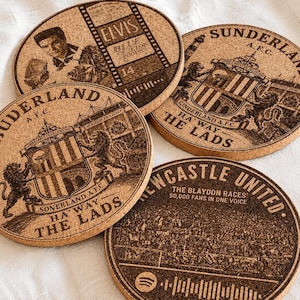 May include: Four round cork coasters featuring dark brown laser-engraved designs. Two coasters display the Sunderland A.F.C. crest with the text 'HA'WAY THE LADS'. One coaster features Elvis Presley with 'BLUE SUEDE SHOES'. One shows Newcastle United with 'THE BLAYDON RACES'.