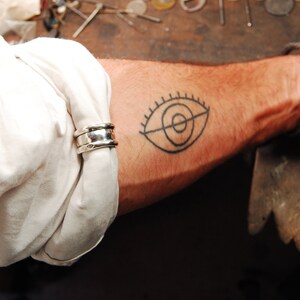 May include: A close-up of a person's forearm with a simple black line drawing tattoo of an eye. The person is wearing a white shirt and a silver ring on their wrist.