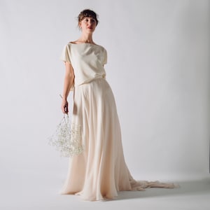 May include: A cream-colored two-piece outfit, featuring a short-sleeved top and a flowing skirt. The skirt has a long train. The outfit is made of a soft, lightweight fabric. The person is holding a small bouquet of white flowers.