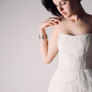 Op de afbeelding: Een vrouw die een witte strapless jurk draagt met delicate kantversiering. Ze draagt een zilveren armband om haar pols.