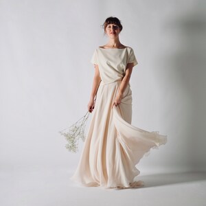 May include: A cream-colored two-piece outfit, featuring a short-sleeved top and a flowing skirt. The skirt is long and drapes elegantly. The outfit is made of a soft, lightweight fabric. The model is holding a small bouquet of white flowers.