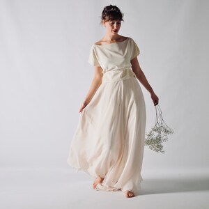 May include: A cream-colored two-piece dress with a boat neckline and short sleeves. The skirt is a flowing, floor-length design. The person is holding a small bouquet of white flowers. The dress is suitable for a wedding or special occasion.