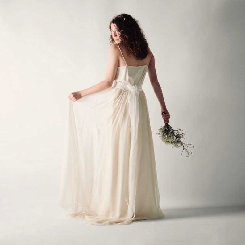 May include: A woman in a white wedding dress with a flowing skirt and a delicate floral crown. She is holding a bouquet of small white flowers.