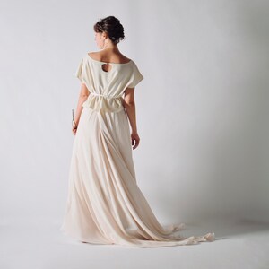 May include: A cream-colored dress with a short-sleeved top and a flowing skirt. The top features a keyhole back and a gathered waist. The skirt drapes elegantly to the floor, creating a soft, romantic silhouette. The dress is made of a lightweight fabric.