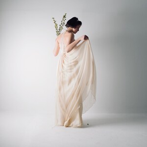 May include: A woman in a flowing, light beige dress with a gathered bodice and a draped skirt. She is holding a sprig of greenery in her hand and is looking down at the dress.